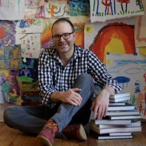 Aaron Slodounik leaning on books in front of a wall covered with children's art